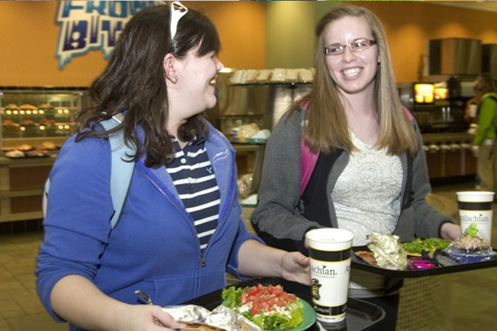 Two people carrying their food and drinks on trays while walking and talking.